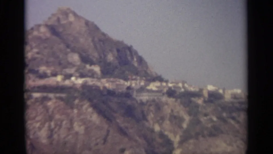 8mm landscape frame with a prominent rocky peak, distant pale buildings, and hazy sky.