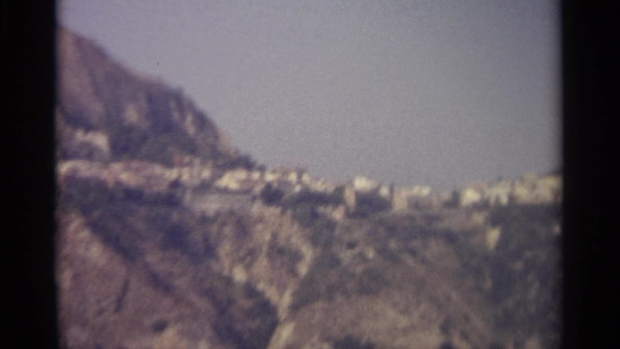 1965 8mm view of a pale hillside town in Sicily, seen across rugged mountain terrain under bright daylight.