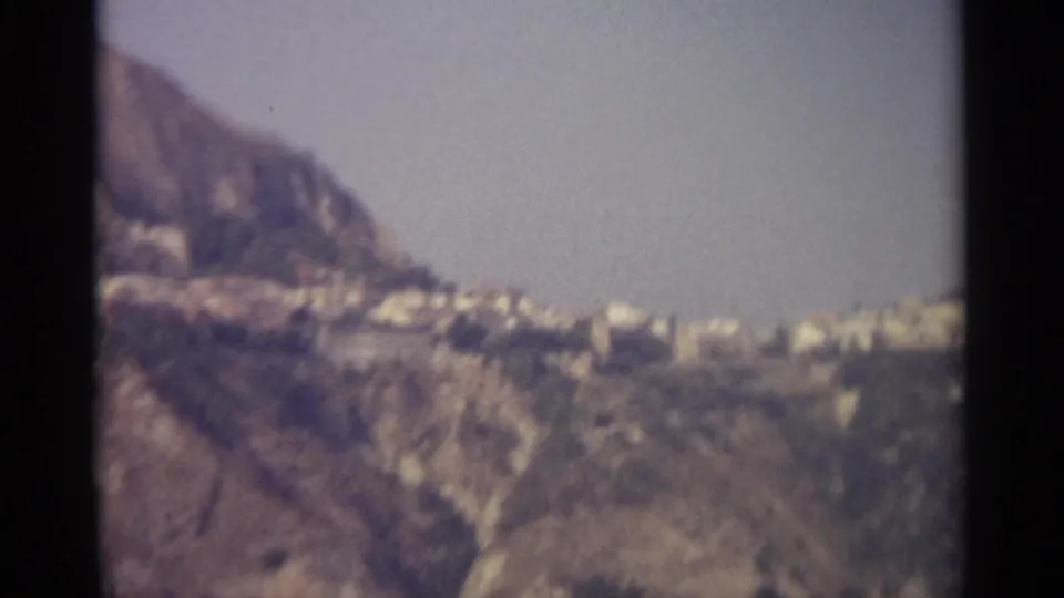 1965 8mm view of a pale hillside town in Sicily, seen across rugged mountain terrain under bright daylight.