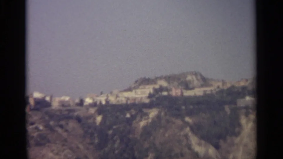 Distant 1965 travel view of a hillside settlement in Sicily, with white buildings set against a broad rocky slope.