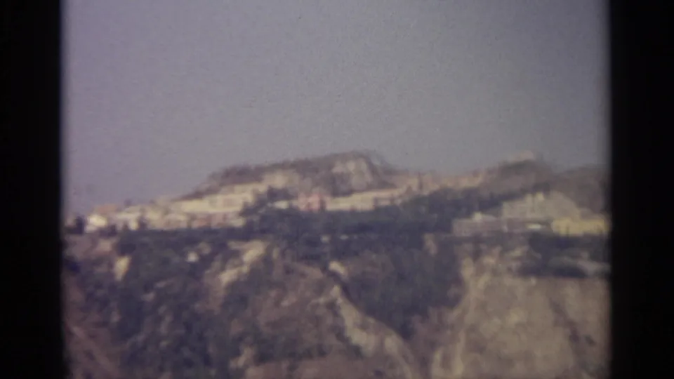 Faded 8mm view of a Sicilian ridge town in bright daylight, with steep brown hillsides below and a clear sky above.