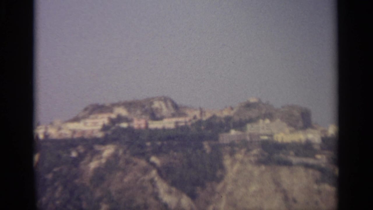 1965 8mm view of a Sicilian hillside town with pale buildings on rugged terrain under a muted sky.
