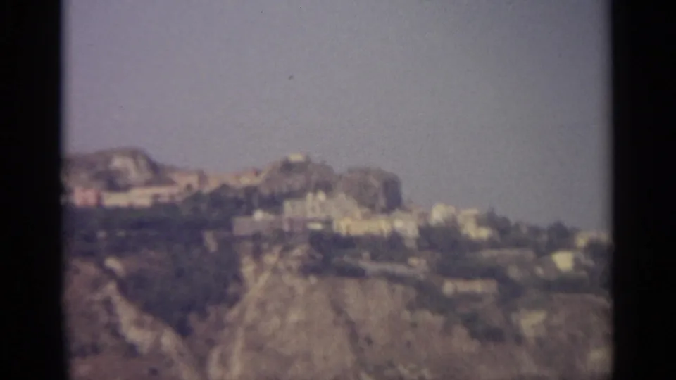 Distant 8mm frame of Sicilian hilltop buildings and rough terrain, softened by grain and faded color.