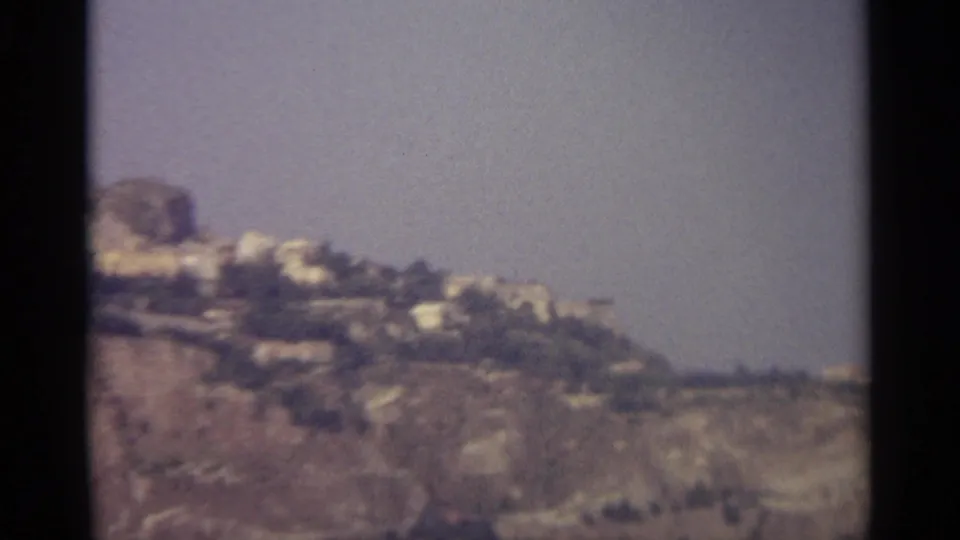 1965 home-movie view of a Sicilian hillside with scattered light buildings and broad pale sky.