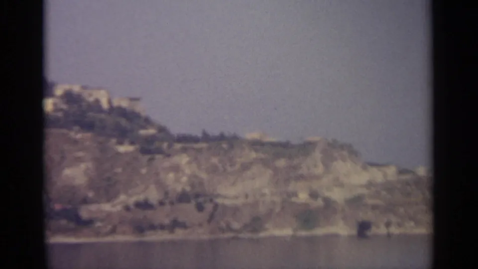 Distant Sicilian hillside view on 8mm film, with ridge-top buildings and possible shoreline below.