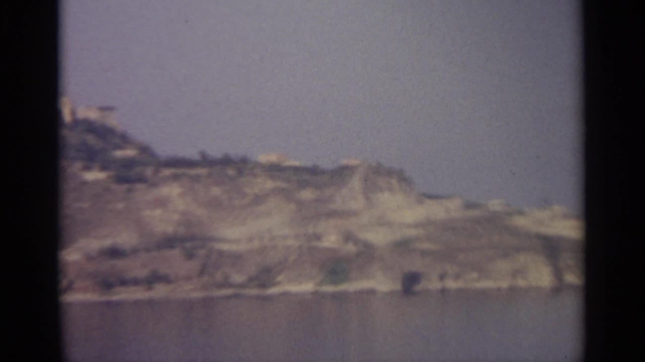 A soft 8mm view of pale coastal cliffs across calm water, likely from a 1965 Mediterranean travel reel.