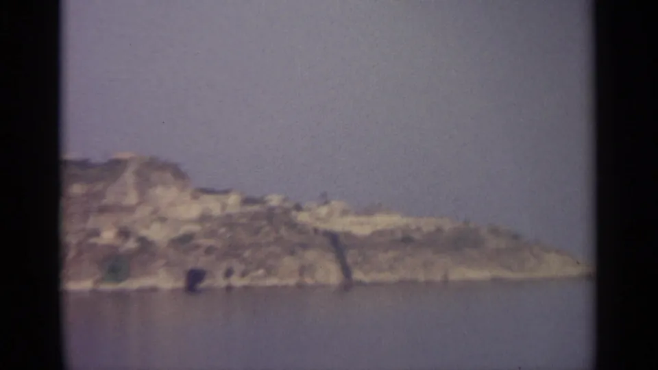 The camera frames a rugged light-toned shoreline across the water, with a hazy sky and muted 8mm color.