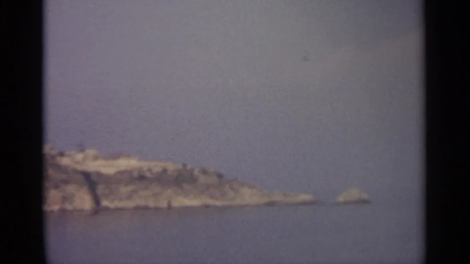 A low section of rocky coastline fades into open water, with a small offshore rock visible through haze.