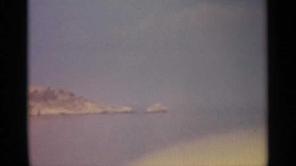 A distant rocky shoreline recedes at left while a small outcrop sits offshore in a soft, hazy 8mm frame.