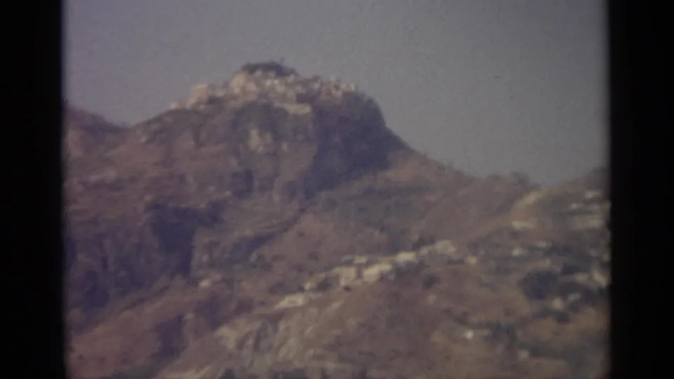 1965 Sicily home-movie frame showing a rocky hill and distant white buildings on dry slopes under a hazy sky.