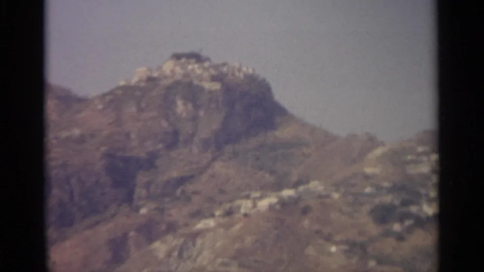 Soft 8mm view of a rugged Sicilian hillside with pale buildings clinging to the slope below a rocky peak.