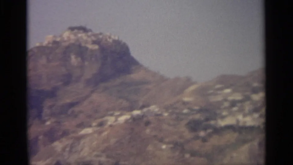 A closer 1965 travel-film frame shows the rocky hill and scattered white hillside buildings in soft focus.