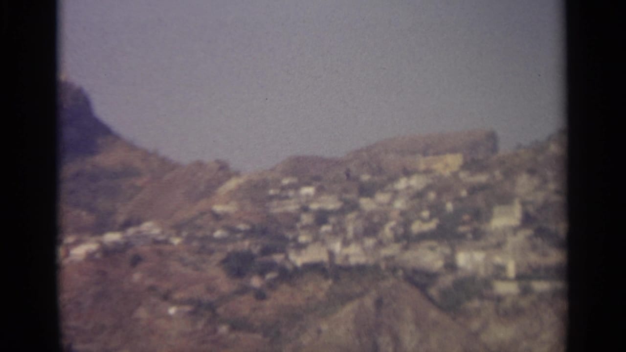 1965 8mm view of a distant mountainside settlement in Sicily, with white buildings clustered across dry slopes.