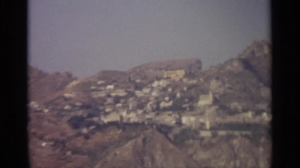 A soft 8mm frame shows a Sicilian mountainside town with pale buildings set against rugged ridges and dry terrain.