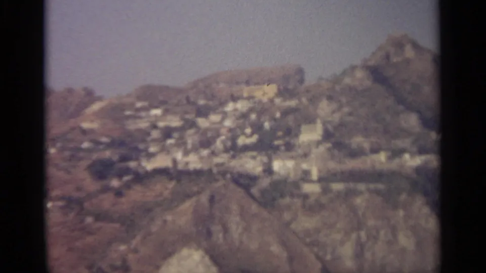 The camera angle shifts across the same Sicilian hillside settlement, revealing more rocky slope and a pointed ridge.