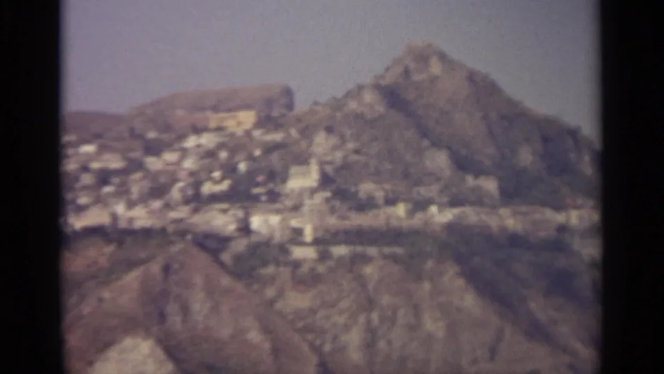 Final 1965 8mm frame of a Sicilian mountain settlement, with white buildings below a jagged ridge and arid slopes.
