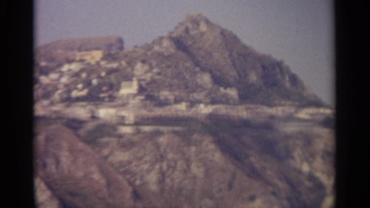 Rocky hillside with pale buildings and hazy sky, framed by dark 8mm film edges.