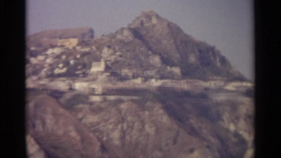 1965 8mm Sicily travel view of a rugged hillside with pale buildings near the slope and a hazy sky.