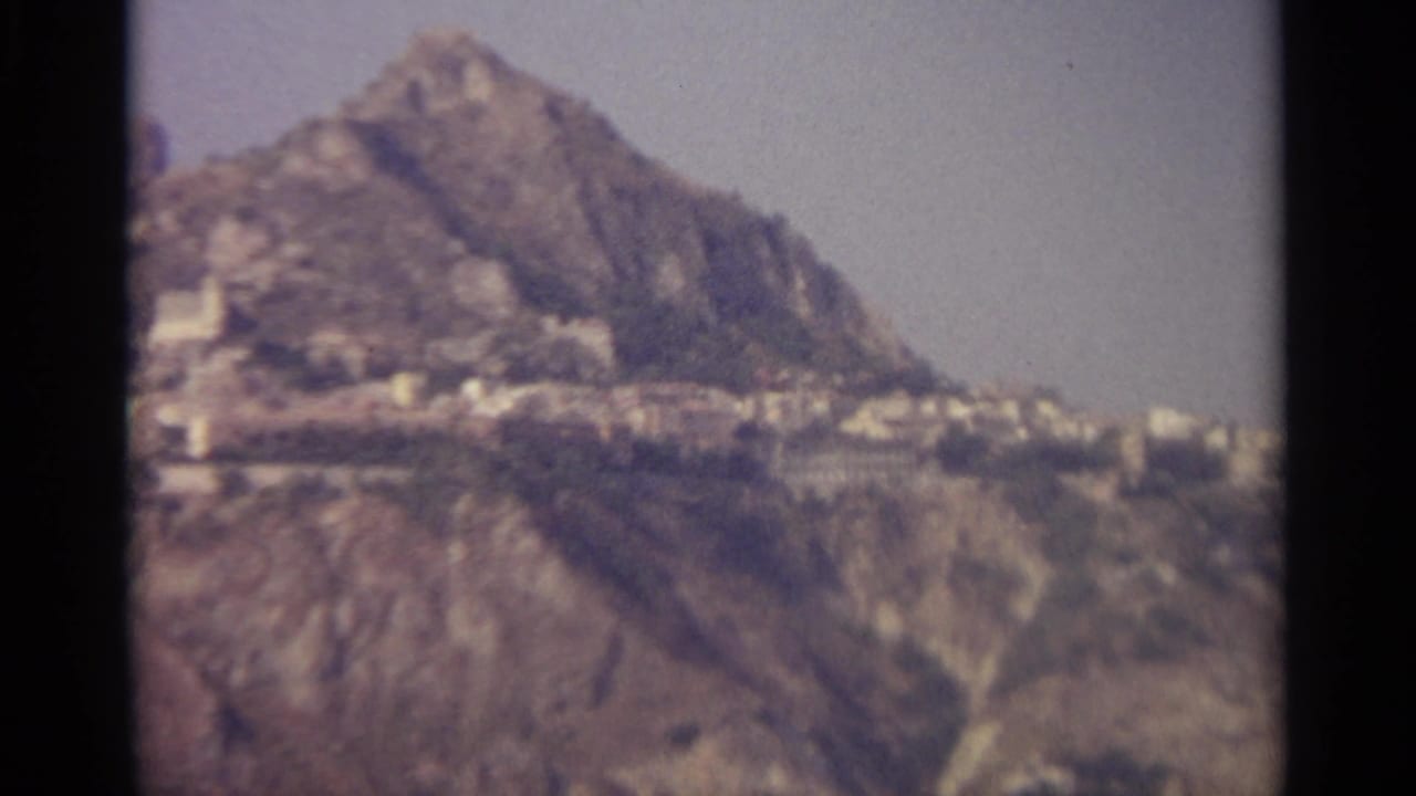 Rugged mountain slope with clustered light buildings below a hazy sky.