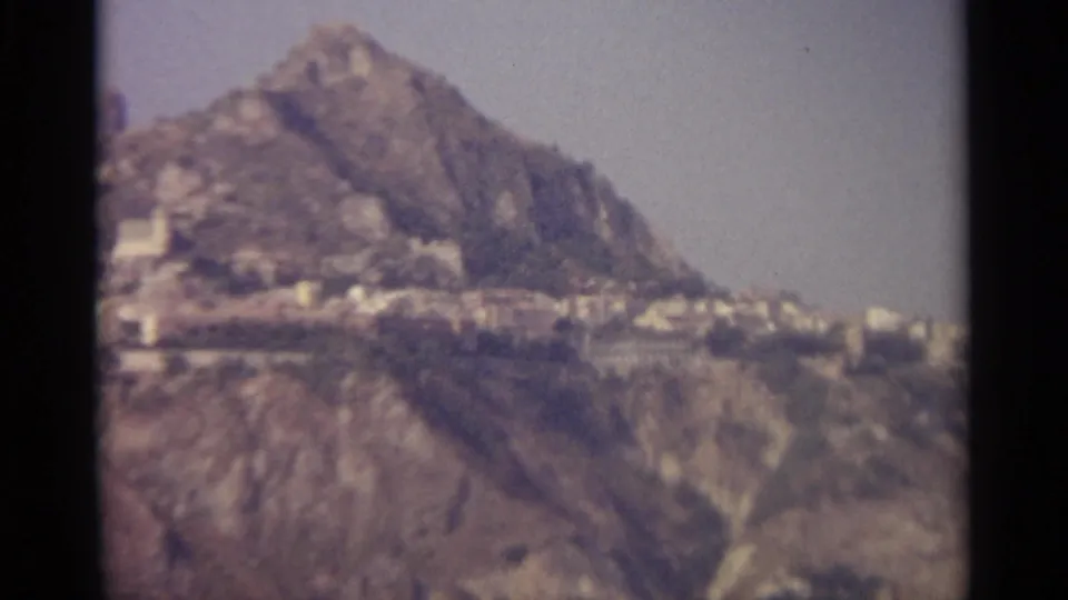 1965 8mm view in Sicily showing a rocky slope and clustered pale hillside buildings in a distant landscape.