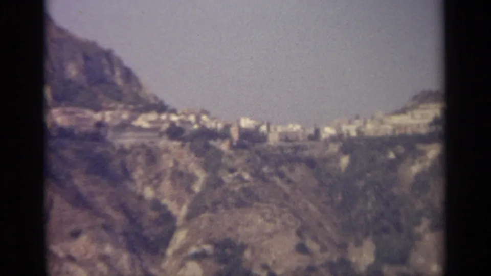 A 1965 Sicily home-movie frame shows distant pale buildings set between rugged slopes and soft, grainy film edges.