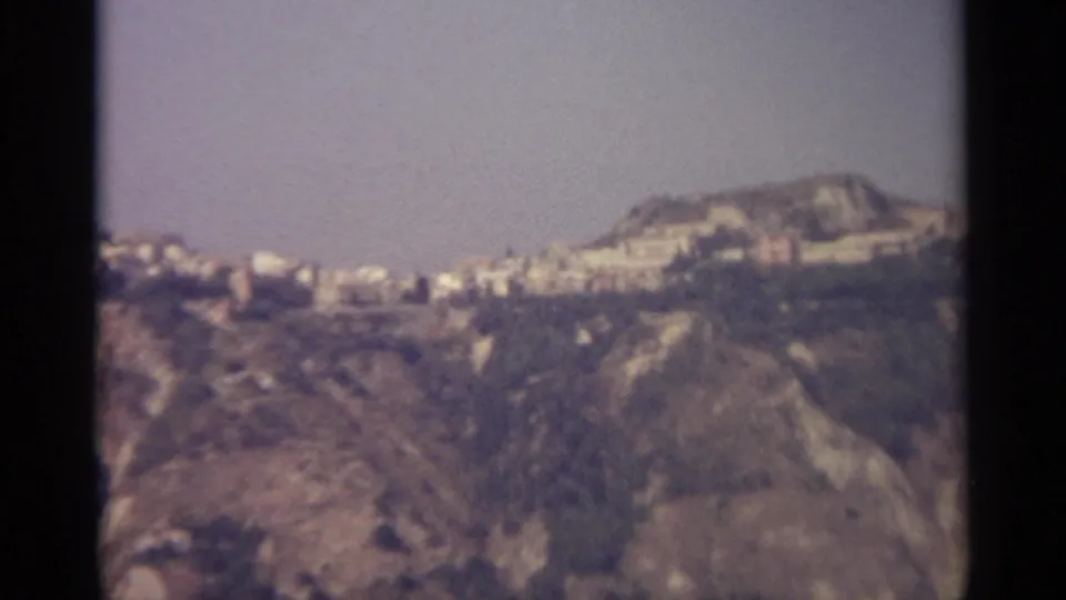 Late-frame 1965 8mm Sicily view of pale hillside buildings and rugged foreground slopes under a muted sky.