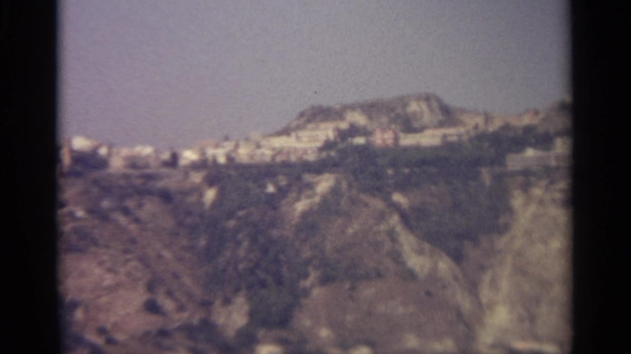 Distant rocky hillside with pale buildings above a calm body of water.