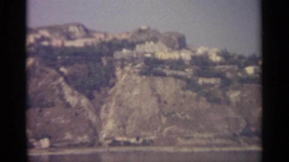 Soft 1965 home-movie frame showing a steep Sicilian hillside with clustered pale buildings over water.