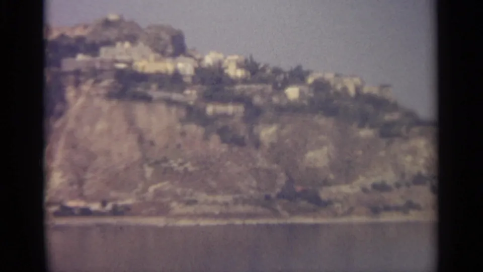 Wide 8mm view of a Sicilian hillside ridge with scattered pale buildings above a calm body of water.