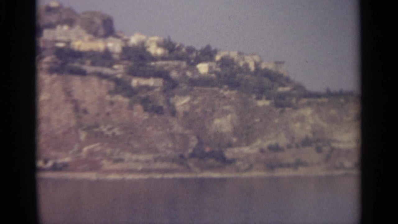 1965 8mm home-movie frame of a rugged Sicily-associated coastline, with distant hillside buildings above still water.