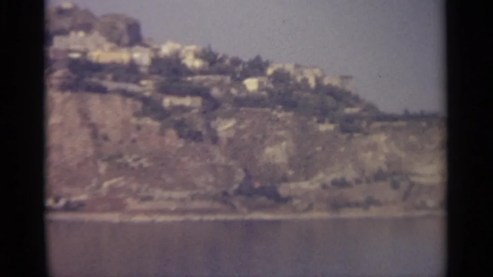 1965 8mm home-movie frame of a rugged Sicily-associated coastline, with distant hillside buildings above still water.