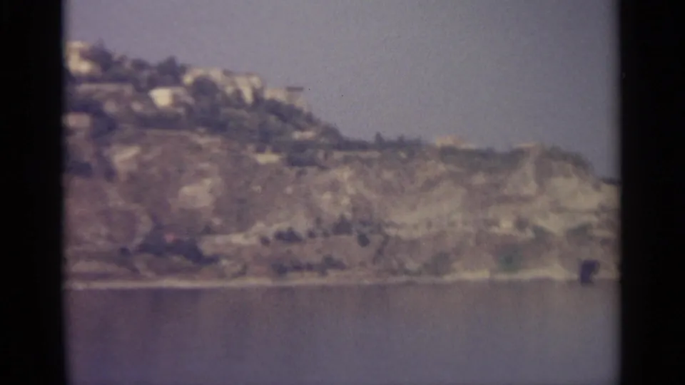 Soft-focus 8mm view of a rugged coastal slope, scattered light buildings, sparse greenery, and calm sea.