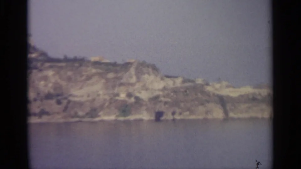Vintage 8mm frame showing a pale rocky shoreline, low vegetation, and calm water in a Sicily-associated coastal view.