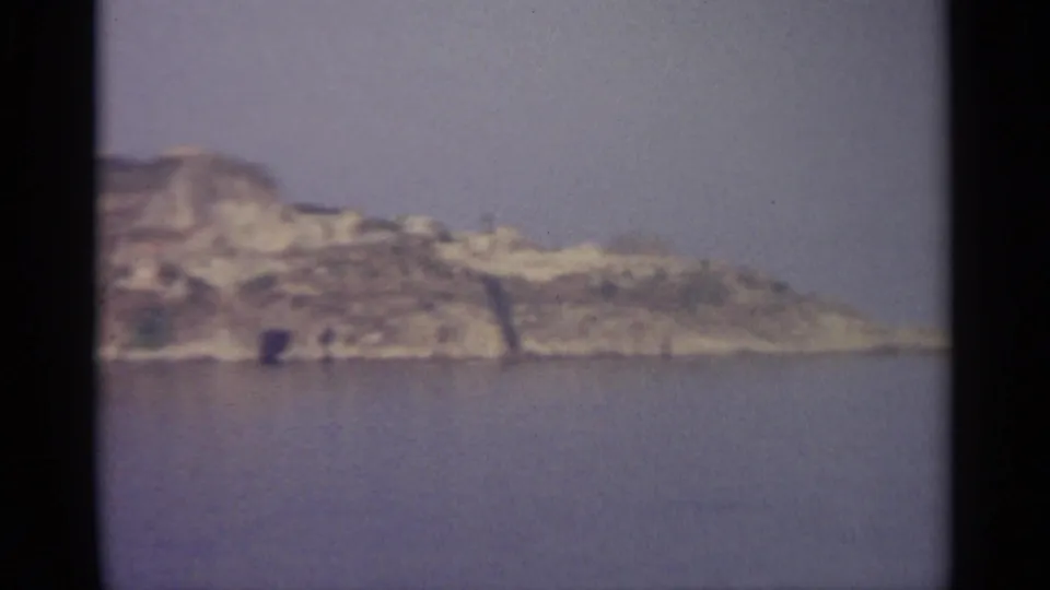 Soft 8mm frame of a rocky coastal headland, still water, sparse greenery, and distant ridge-top structures.