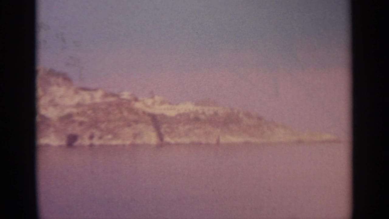Rocky island across calm water under a clear sky, framed by dark film edges.