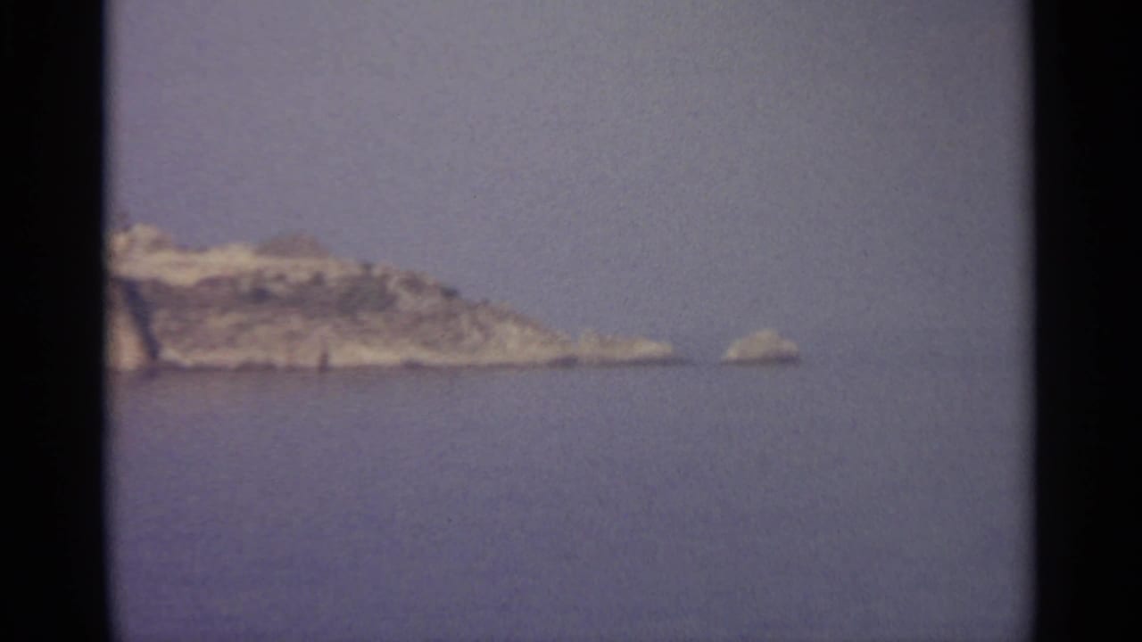 Rocky headland and small offshore rock seen across calm blue water.