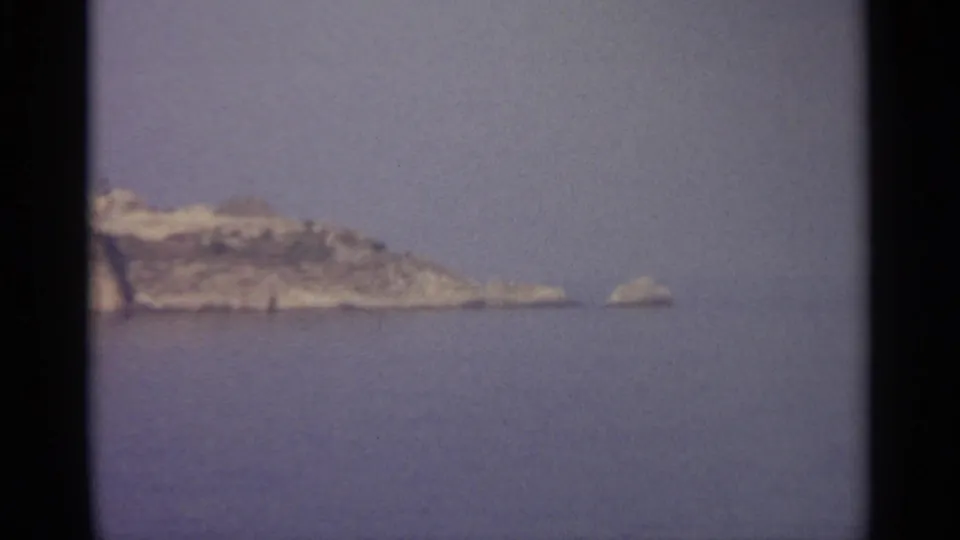 A rocky island or headland appears in the distance across calm Mediterranean-looking water, with grain and dark 8mm frame borders.