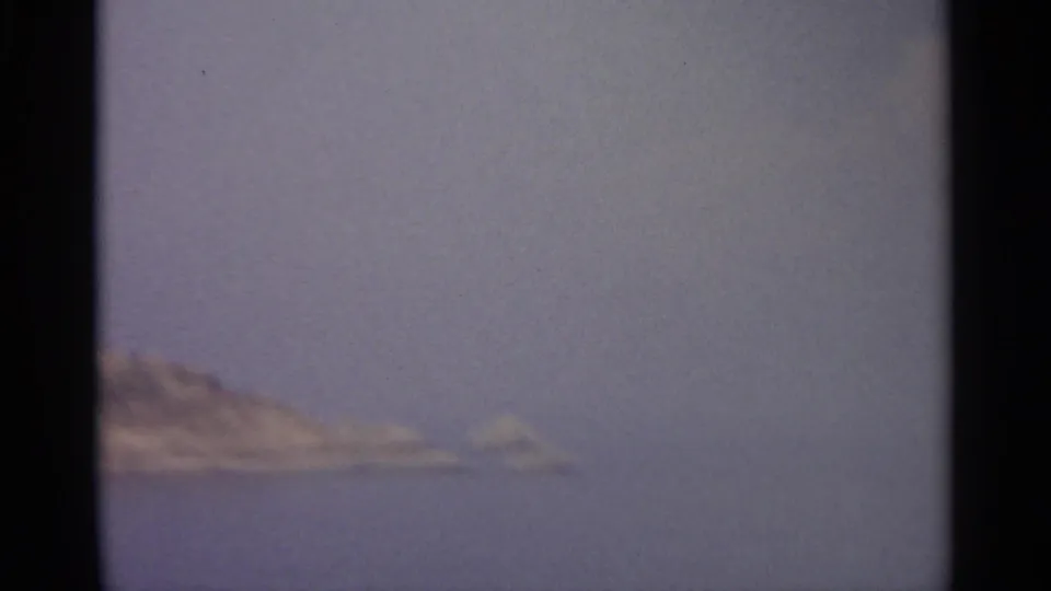 A rocky coastal edge appears low in the frame, surrounded by calm blue water and a pale sky in soft 1960s 8mm color.