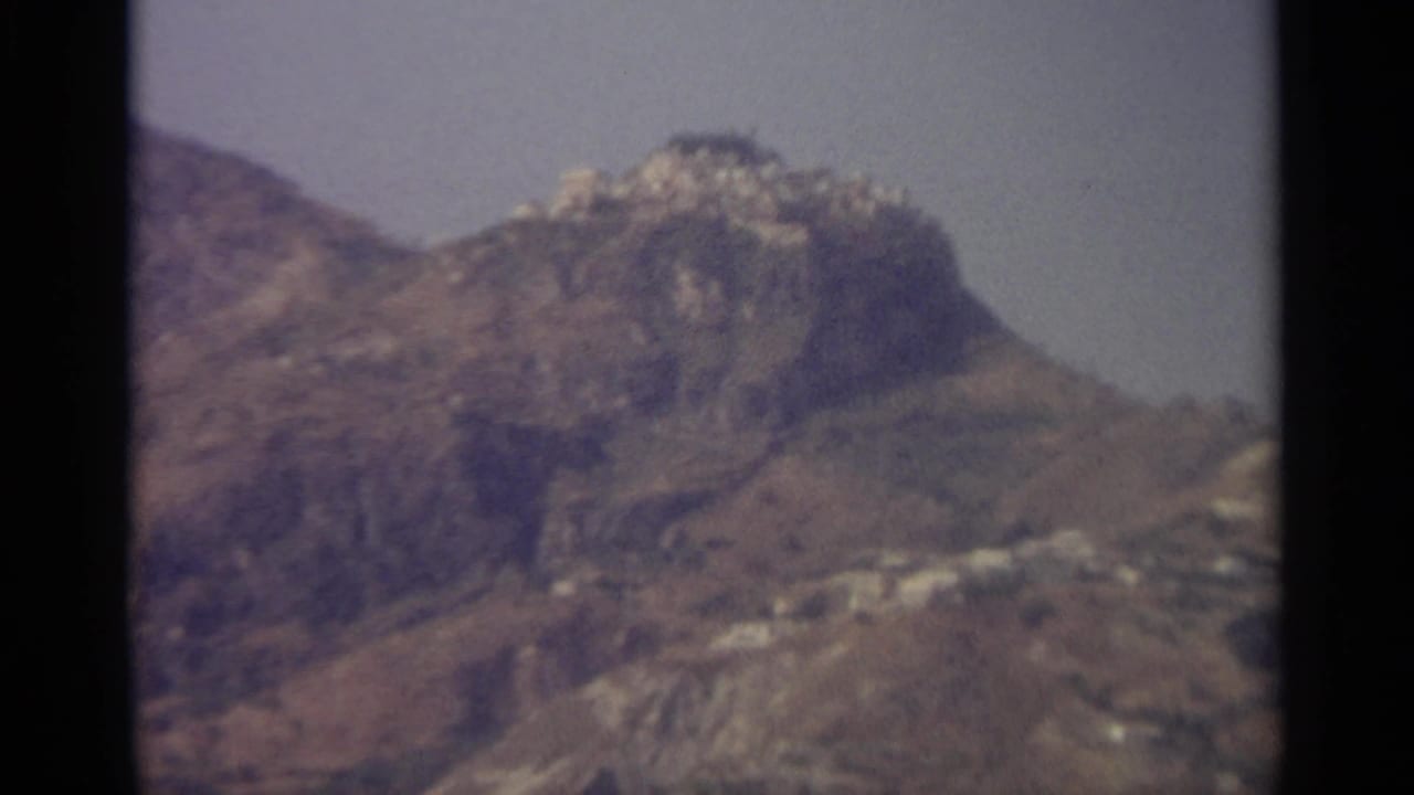 Soft 8mm frame of a rocky Sicilian hilltop landscape with a small cluster of pale buildings visible along the ridge.