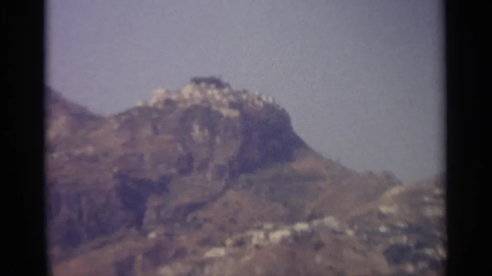 Blurred 8mm frame of a Sicilian mountain peak, dry slopes, and small pale buildings spread below the ridge line.