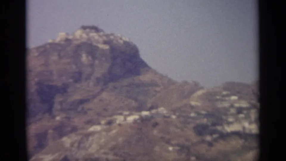 End frame of the 8mm pan showing a craggy Sicilian hilltop and pale hillside buildings against a hazy sky.