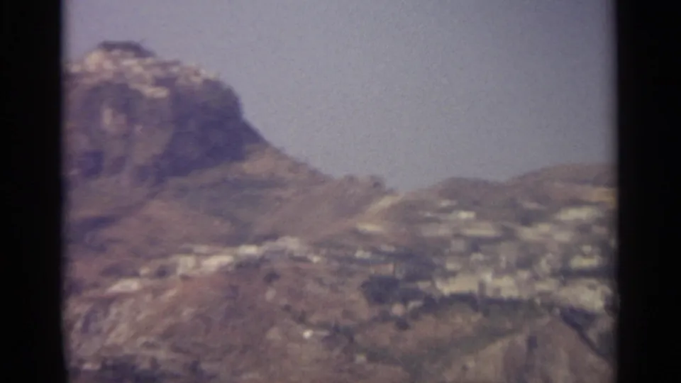 1965 8mm view of a Sicilian hillside settlement with rugged mountains and pale buildings in soft focus.