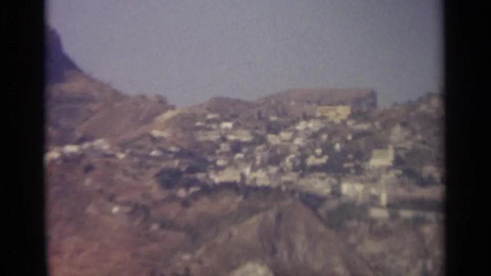 Distant Sicilian mountain village appears across rocky slopes, with dark frame borders from the 8mm scan.