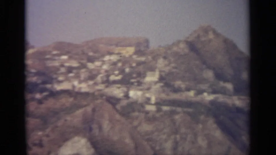 1965 rural Italy home-movie frame showing a pale hillside town below rugged Sicilian mountains.