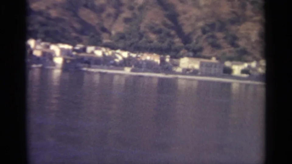 A Sicilian lakeshore settlement appears below wooded mountains, captured in faded 1965 8mm color.