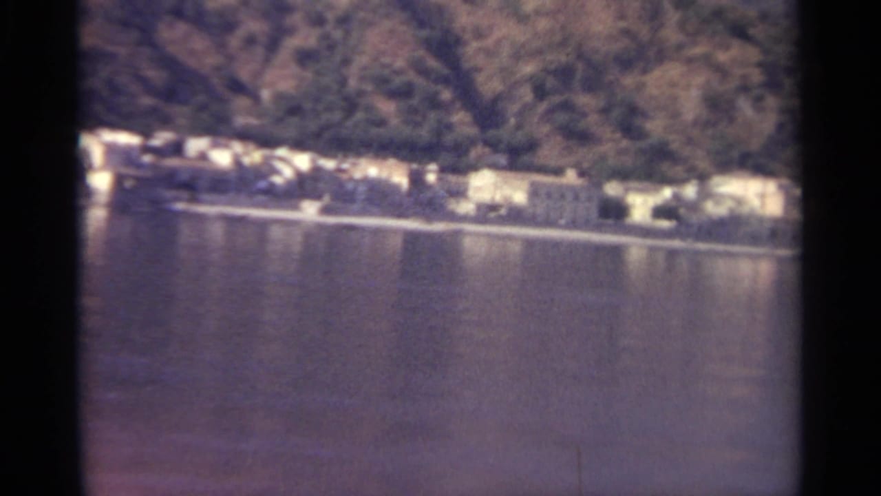 Distant shoreline buildings across calm lake water below a wooded hillside.