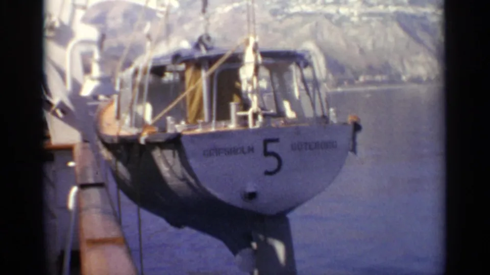 A small boat labeled “GRIPSHOLM 5 GÖTEBORG” hangs from dockside crane lines beside calm water.