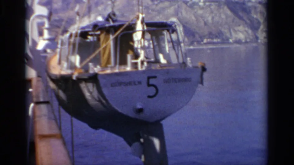 The small Gripsholm sailboat appears suspended beside the ship, with the hull number 5, lifting ropes, blue water, and shoreline visible.