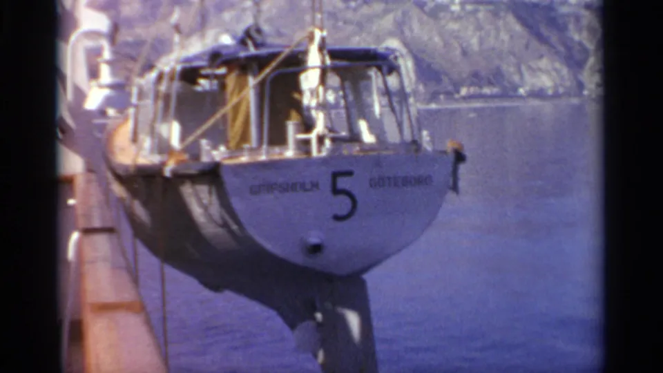 A small sailboat with visible hull lettering and number 5 remains suspended from lines beside a larger vessel in archival 8mm footage.