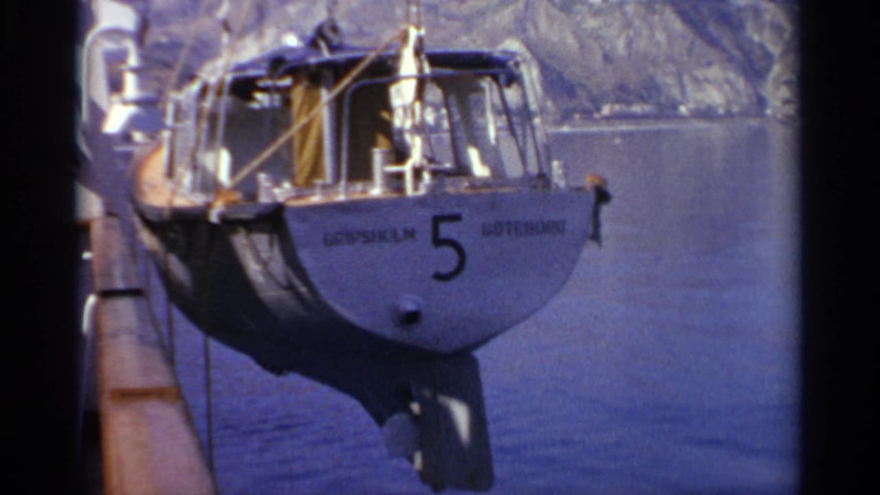 1965 8mm frame showing a grey boat marked 5 at a pier, with calm water and a rocky coastal hillside in the background.